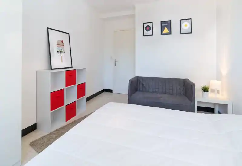 Chambre lumineuse et minimaliste avec une couette blanche au premier plan, un petit canapé gris et une étagère avec boîtes de rangement rouges — espace calme et bien éclairé, parfait pour un séjour en ville.