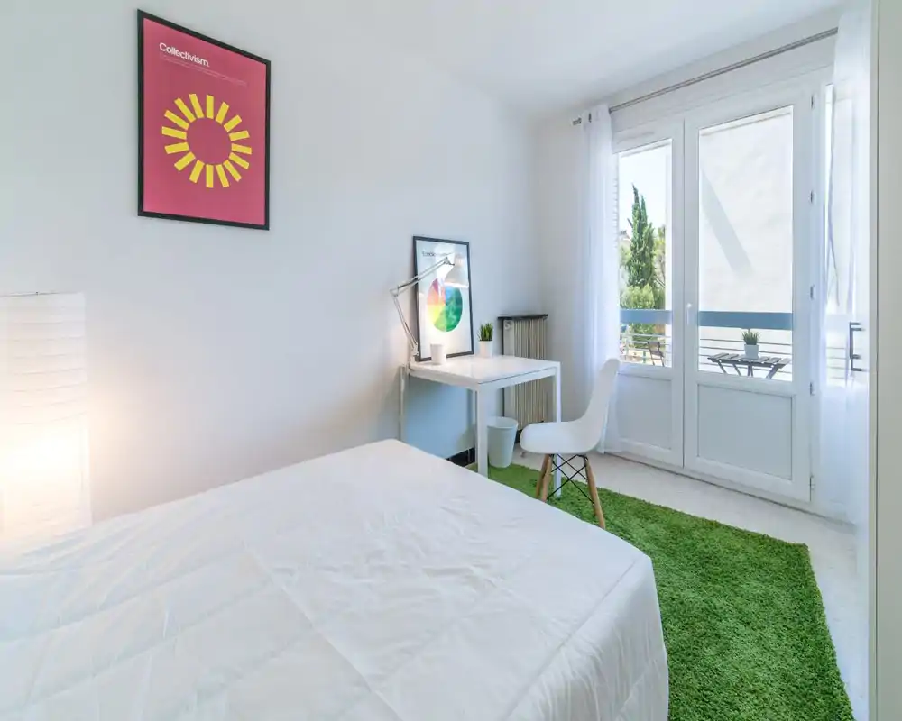 Chambre lumineuse et minimaliste avec lit blanc, bureau et grandes fenêtres donnant sur un petit balcon — moderne, aérée et bien éclairée pour une annonce.