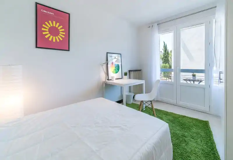 Chambre lumineuse et minimaliste avec lit blanc, bureau et grandes fenêtres donnant sur un petit balcon — moderne, aérée et bien éclairée pour une annonce.