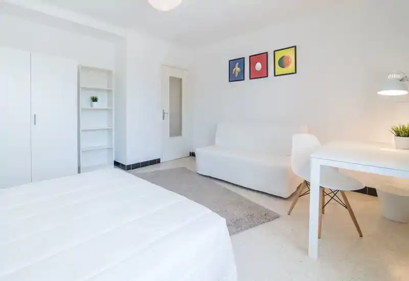 Bright, minimalist bedroom with a visible bed in the foreground, a white sofa and a simple desk — ideal for a clean, modern rental listing.