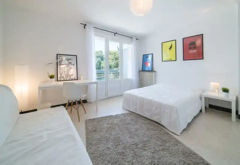Bright, minimalist bedroom with a queen-size bed, soft rug and a workspace by large windows that overlook greenery. The room feels airy and clean with modern wall art and thoughtful bedside lighting.