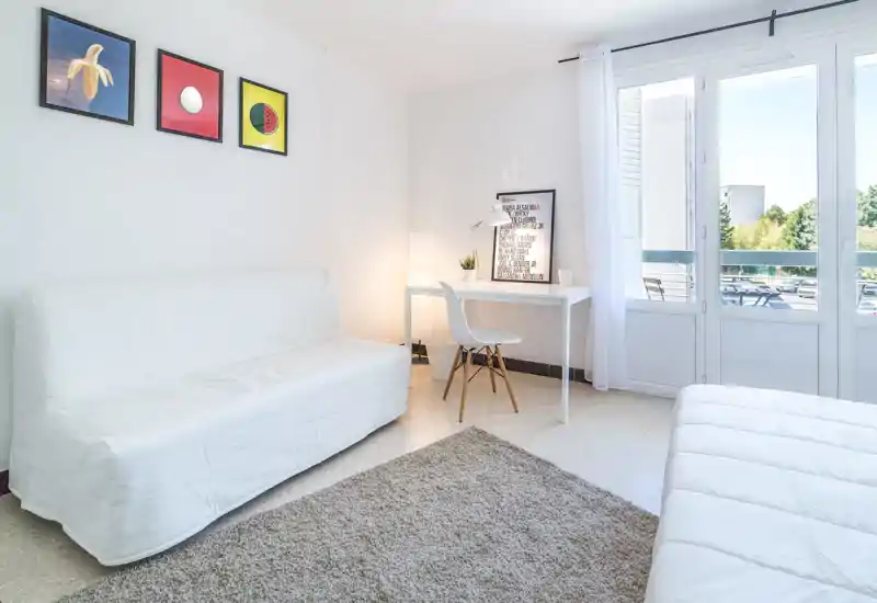 Bright, minimalist bedroom with a visible white mattress in the foreground, a sofa, desk and large windows opening to a balcony; neutral decor and natural light create an airy, calm space.