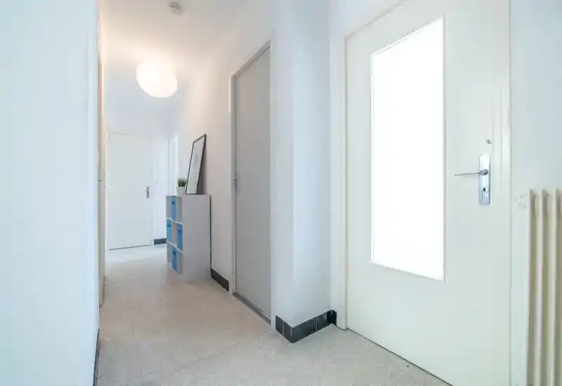 Entrée lumineuse et minimaliste avec portes blanches, sol carrelé et une petite étagère offrant rangement et décoration.