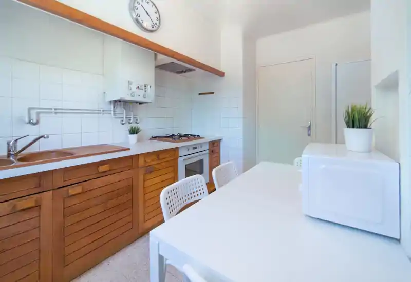 Cuisine compacte et lumineuse avec placards bas en bois, plaque à gaz et four encastré. Plans de travail blancs et petite table avec chaises pour des repas pratiques au quotidien.