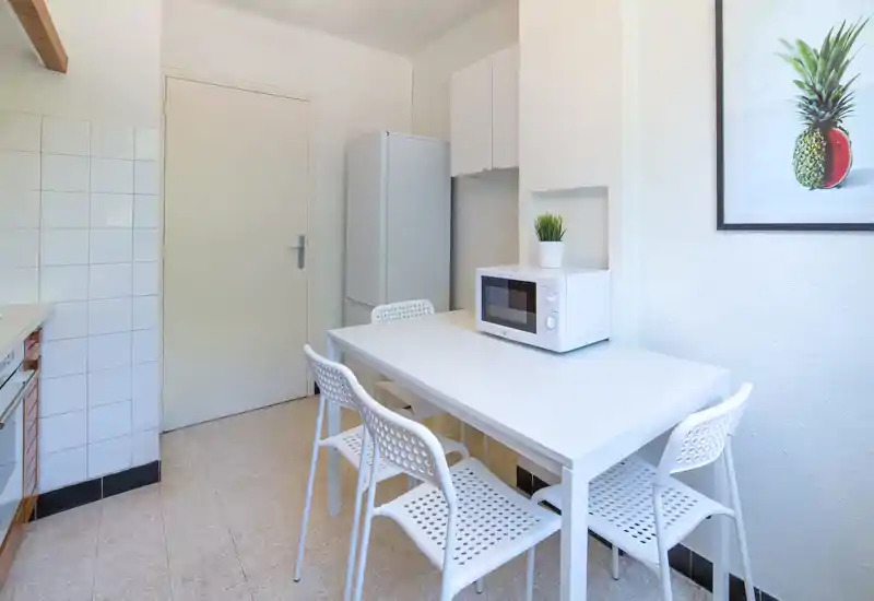 Cuisine compacte et lumineuse avec une table blanche, quatre chaises perforées, un micro-ondes et un grand réfrigérateur ; décoration minimaliste et style épuré.