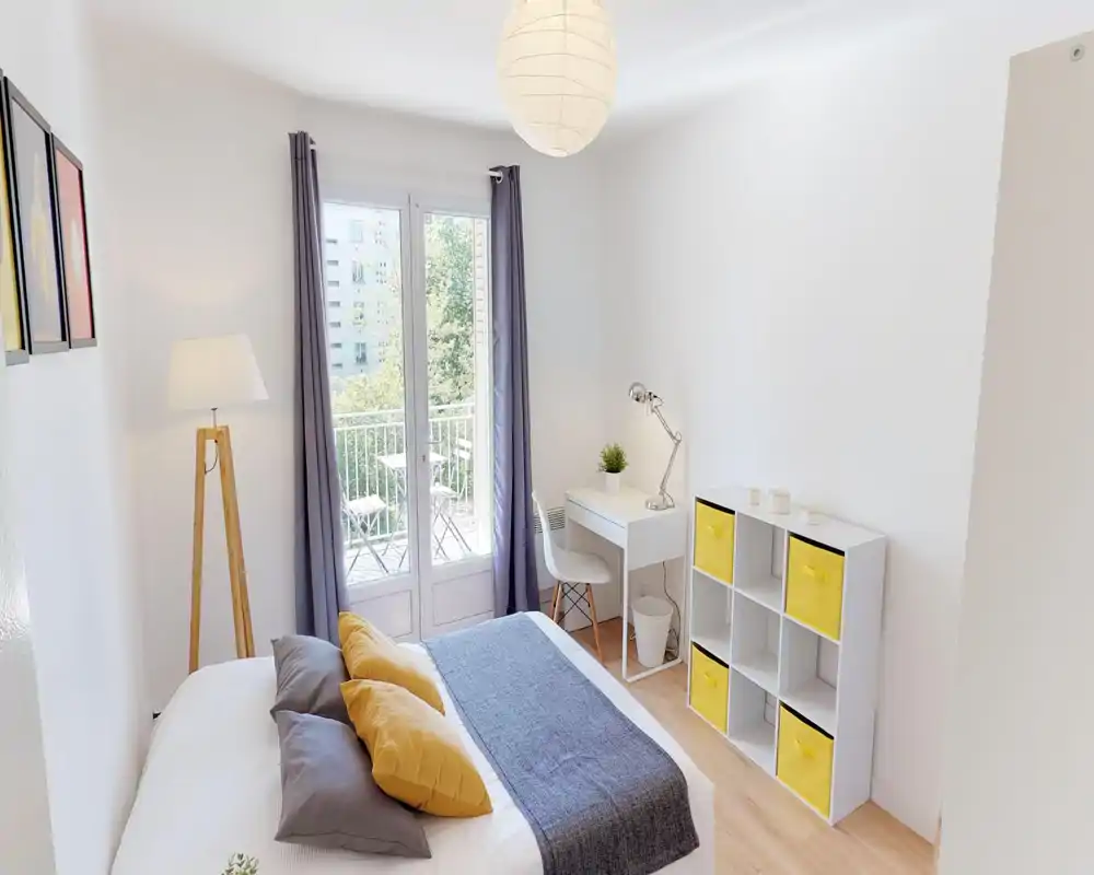 Camera da letto luminosa e moderna con letto matrimoniale ben rifatto, cuscini accoglienti e un runner. Piccola scrivania vicino alla finestra e scaffale con contenitori gialli che completano l’aspetto minimal e arioso con accesso al balcone.