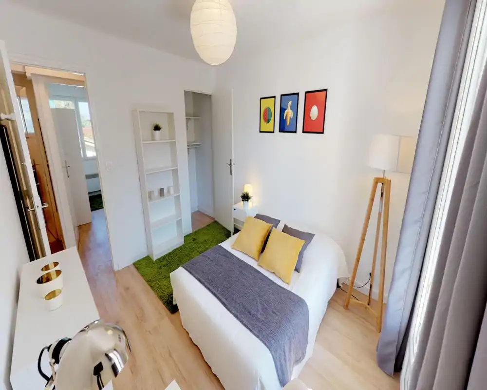Camera da letto luminosa e moderna con letto matrimoniale in lenzuola bianche, runner grigio e cuscini gialli; una lampada da terra, comodino e scaffale rendono lo spazio accogliente e ben illuminato.