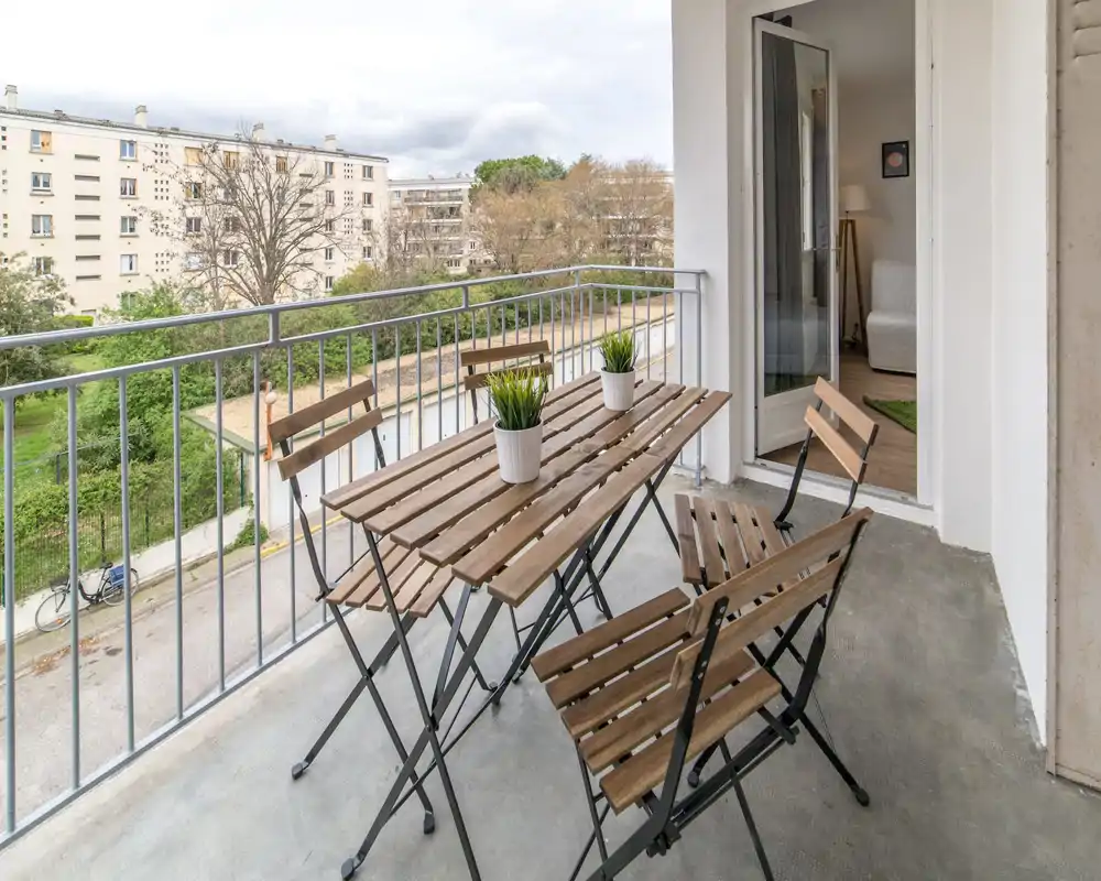 Balcone soleggiato con tavolo bistrò in legno e quattro sedie pieghevoli, due vasetti con piante e vista su cortili verdi e palazzi vicini.