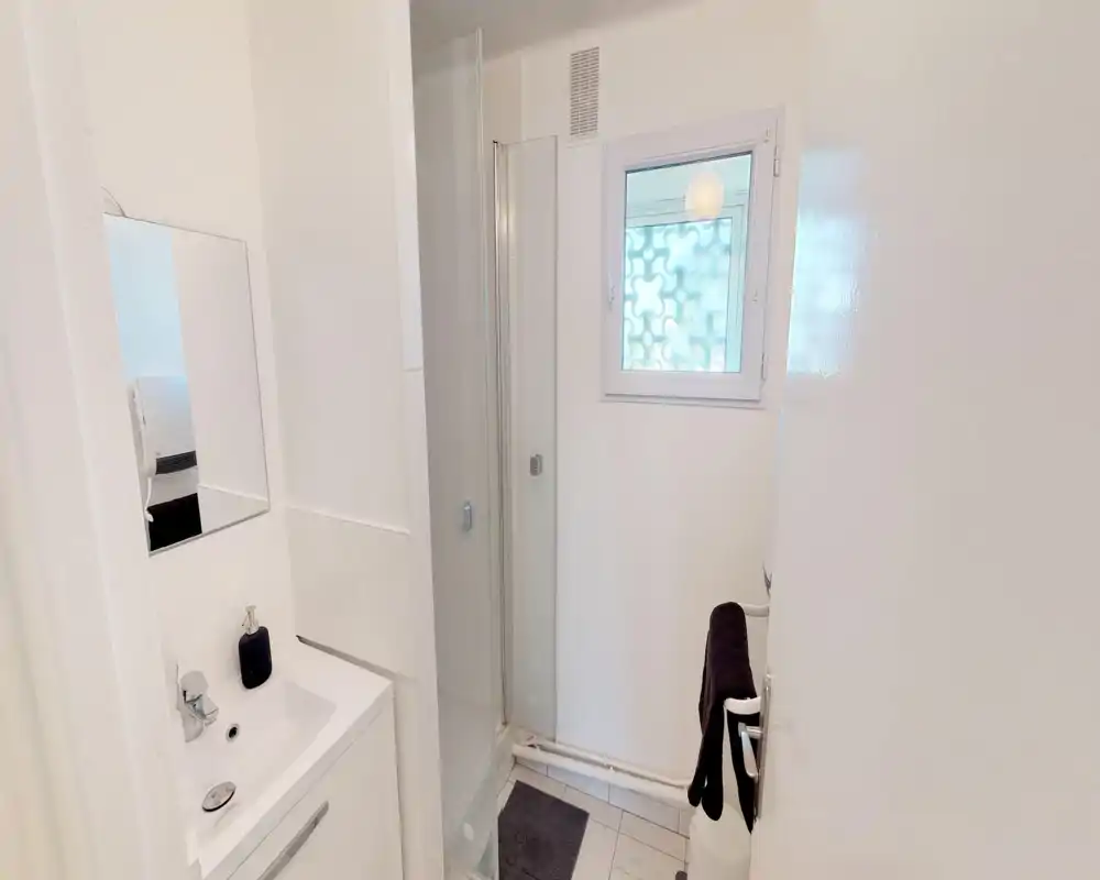 Bagno compatto e luminoso con cabina doccia, lavabo e finestra. Piastrelle bianche e mobile bagno moderno e semplice.