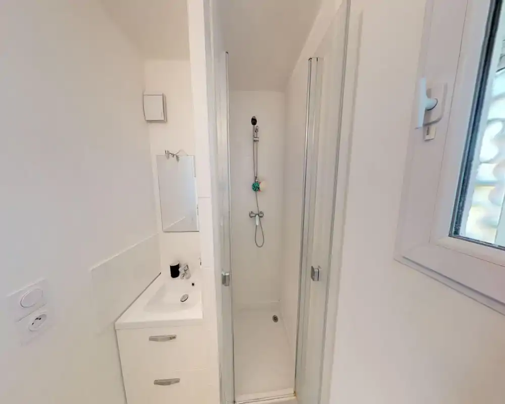Bagno compatto tutto bianco con doccia a filo, porte a vetro richiudibili, piccolo lavabo con mobiletto e una finestra che garantisce luce naturale.