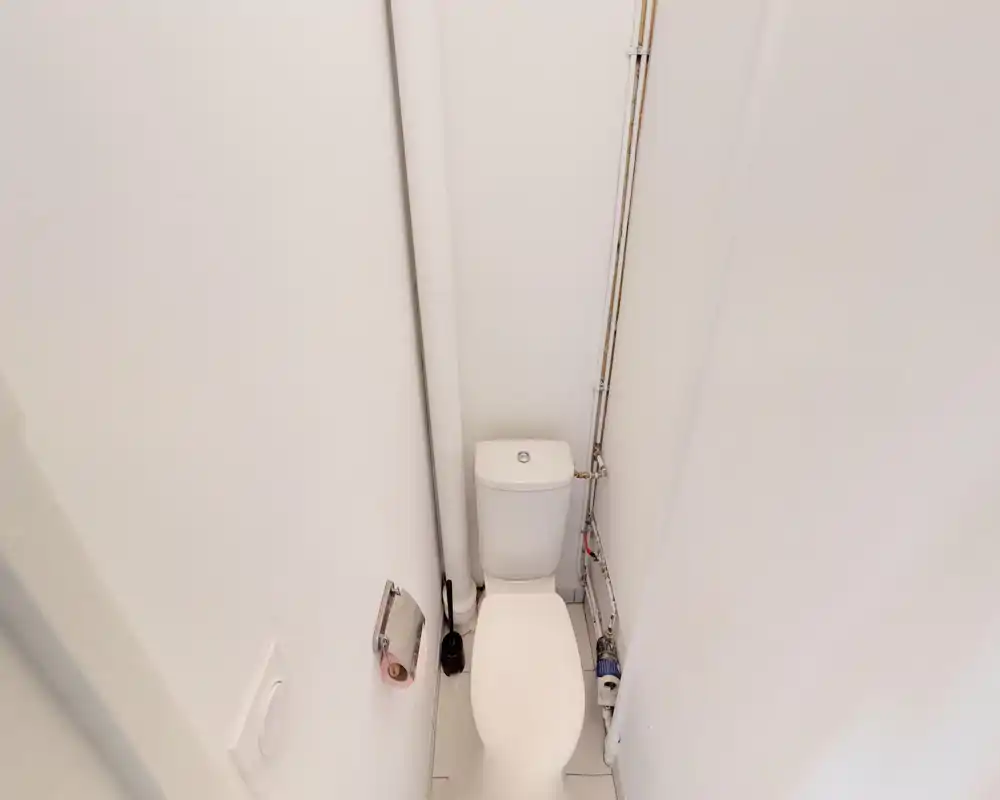 Piccolo bagno con wc e tubazioni a vista, portarotolo a parete e finiture essenziali. Ambiente luminoso e pulito.
