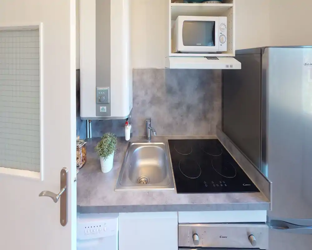 Cucina moderna compatta con lavello in acciaio, piano a induzione, microonde e piccolo frigorifero; piani di lavoro effetto pietra e una pianta decorativa creano uno spazio ordinato e funzionale.
