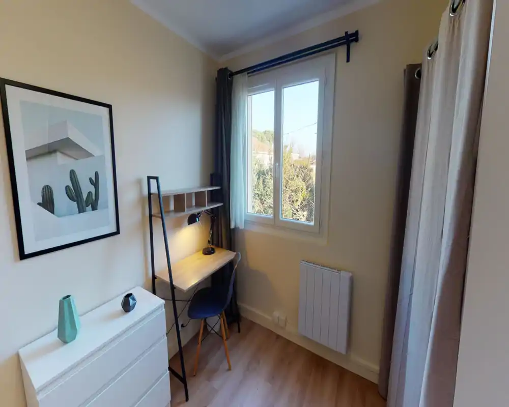 Small bedroom/office corner with a compact desk by a bright window, simple chest of drawers and neutral decor — tidy and well lit.