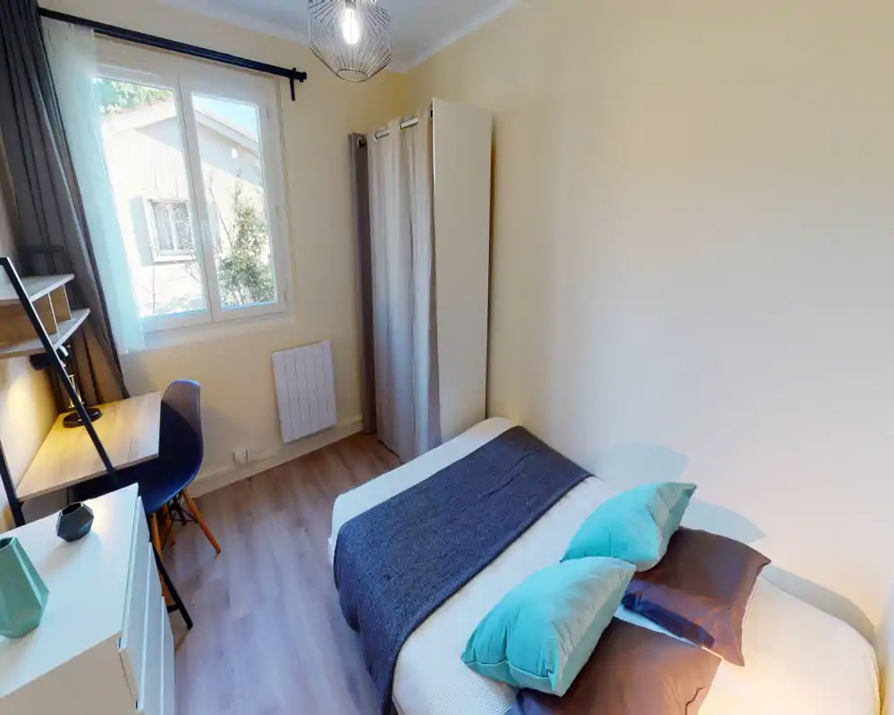 Bright, simply furnished bedroom with a double bed dressed in neutral linen and colorful cushions, a compact desk by the window and a small wardrobe concealed by a curtain — well-lit and neatly staged for a rental listing.