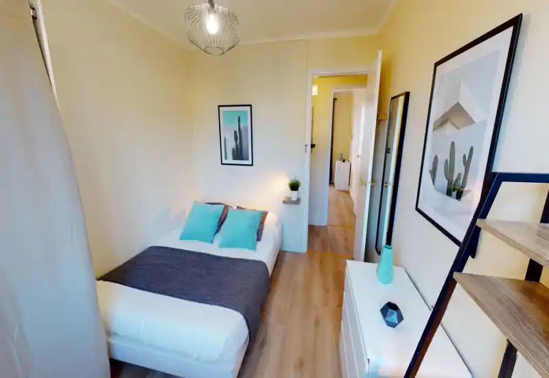 Compact, well-styled bedroom with a double bed dressed in white linens, teal accent pillows and a dark throw. The room is bright, neatly decorated with framed art and a small bedside shelf with a plant, offering a cozy feel ideal for short-term rentals.