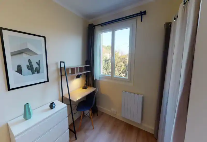 Small bedroom/office corner with a compact desk by a bright window, simple chest of drawers and neutral decor — tidy and well lit.