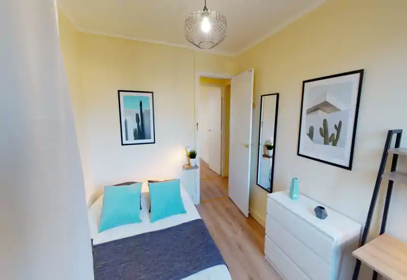 Bright, modern bedroom with a neatly made double bed accented by turquoise cushions, framed wall art, a white dresser and a tall mirror — ideal for a cozy rental listing.