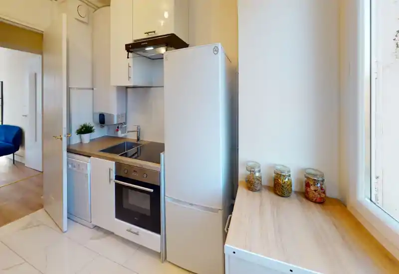 Compact modern kitchen with white cabinets, integrated oven, fridge and countertop jars; bright natural light from a nearby window creates a clean, welcoming workspace.