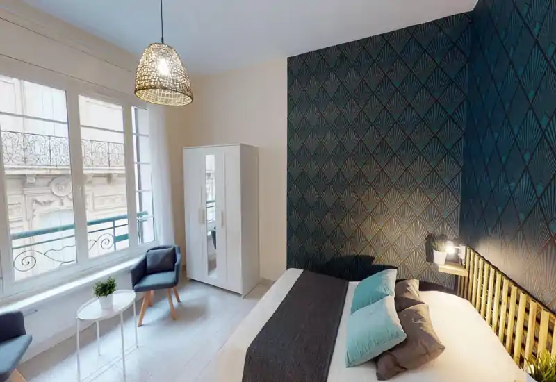 Bright, stylish bedroom with patterned accent wall, a large comfortable bed dressed with cushions and a woven bedside lamp creating a warm atmosphere. Large windows provide abundant natural light and a small seating area with armchairs and a table near the wardrobe.