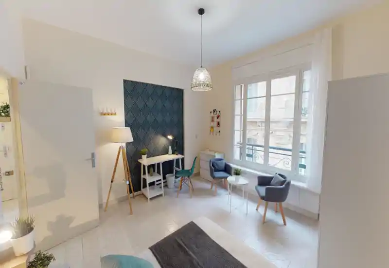 Bright, stylish bedroom with a visible double bed in the foreground, a decorative accent wall, a compact desk and two armchairs by a large window — ideal for both relaxation and light work.