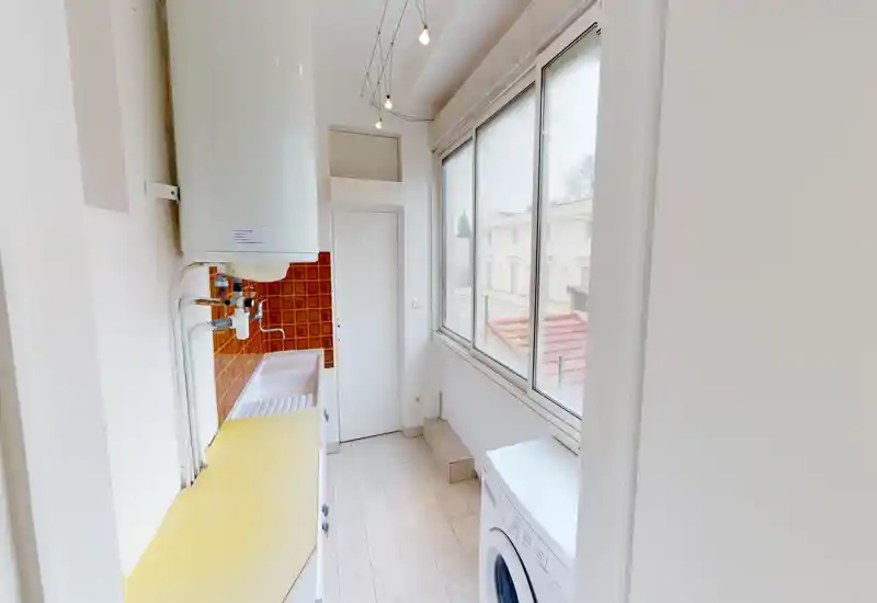 Kitchenette/coin buanderie compact avec plan de travail, évier et lave-linge près de grandes fenêtres offrant une belle lumière naturelle.