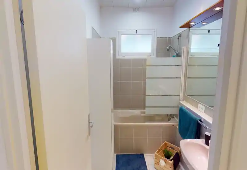Salle de bain propre et lumineuse avec baignoire et paroi de douche, murs carrelés, lavabo avec miroir et petit rangement. Tons neutres et agencement pratique, idéal pour une annonce locative.