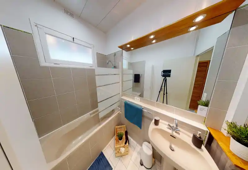 Salle de bain moderne et lumineuse avec baignoire/douche, lavabo, grand miroir et accessoires soignés ; idéal pour mettre en valeur une pièce propre et bien éclairée.