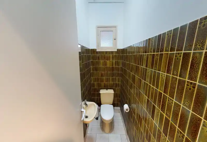 Salle d'eau compacte avec WC et petit lave-main mural, murs carrelés marron-vert décoratifs et fenêtre dépoli apportant de la lumière.