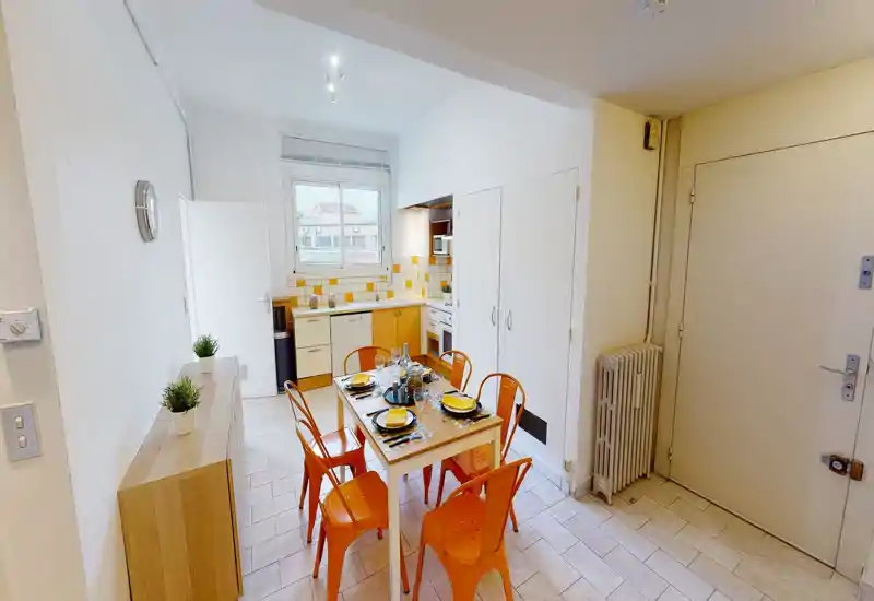 Cuisine compacte avec table à manger pour six, chaises métalliques orange et une fenêtre apportant la lumière naturelle. La crédence carrelée et la table dressée donnent une ambiance chaleureuse, parfaite pour les locations de courte durée.
