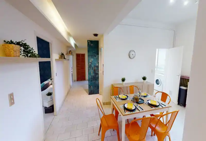 Coin repas lumineux et aménagé avec une table dressée et des chaises orange colorées, idéal pour les repas et petits rassemblements ; l'espace communique avec les pièces adjacentes et la buanderie.