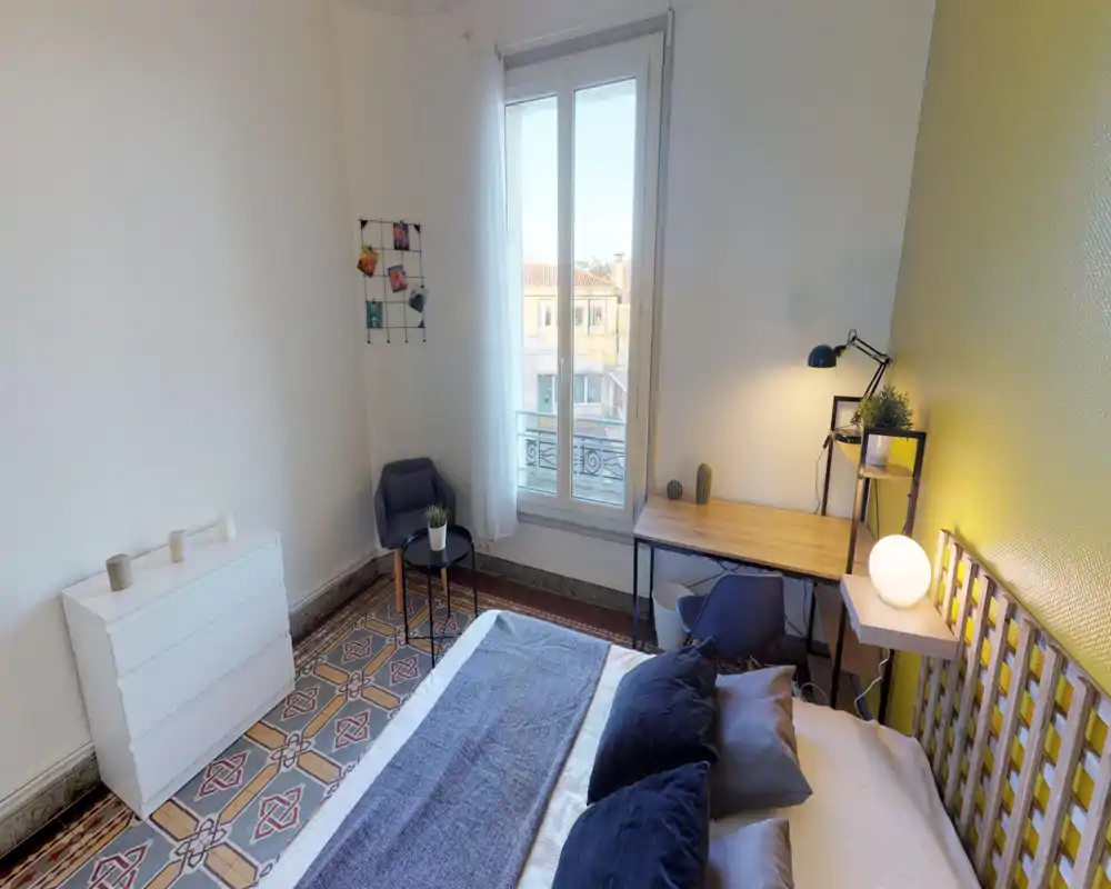 Chambre lumineuse et stylée avec un lit double confortable, coussins décoratifs et une tête de lit en bois. La grande fenêtre apporte de la lumière naturelle sur un bureau et un coin détente.