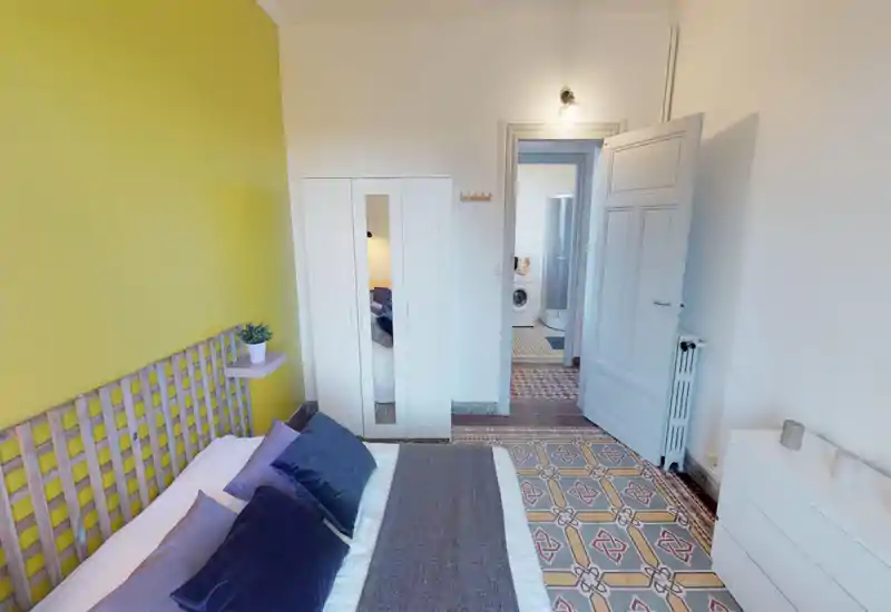 Bright, stylish bedroom with a large bed, decorative pillows, a wooden slatted headboard and patterned tiled floor. The room features a white wardrobe and dresser, plus a small wall shelf with a plant for a cozy touch.