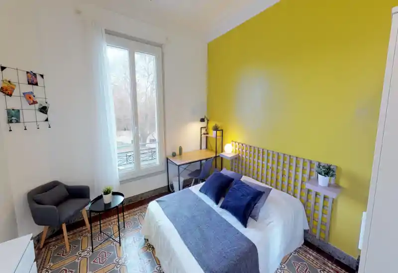 Bright, stylish bedroom with a double bed, blue cushions and a contrasting yellow accent wall. The room features a small desk by the window, a cozy armchair, patterned tile floor and decorative plants—ideal for a comfortable stay.