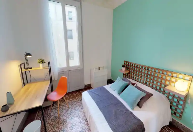 Bright, stylish bedroom with a double bed, decorative pillows and coordinated teal accent wall. A workspace with a wooden desk and orange chair sits by the window, and patterned floor tiles add character.