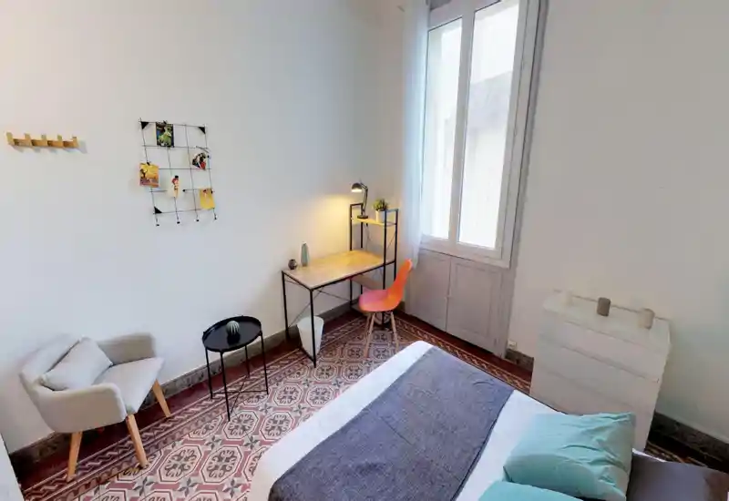 Bright, tidy bedroom with a double bed in the foreground, patterned tiled floor and a small workspace by a large window. The space features a comfortable armchair, decorative textiles and warm desk lighting for a cozy atmosphere.