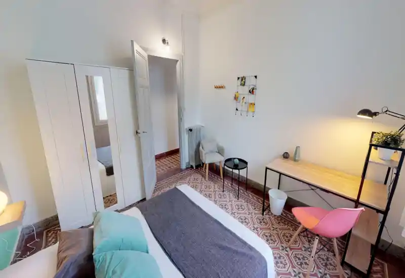 Bright, well-staged bedroom with a double bed in the foreground, patterned tiled floor and a small workspace by the window. The room feels tidy and inviting, with soft textiles and warm lighting.