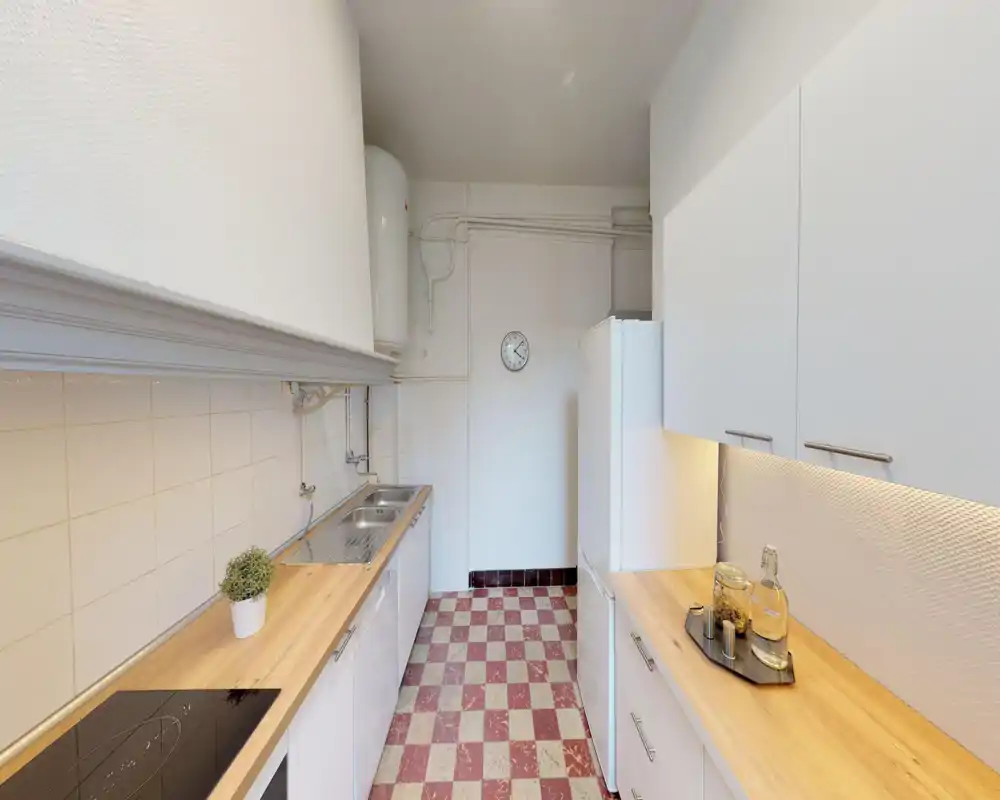 Bright, narrow kitchen with white cabinets, wooden countertops and a checkerboard floor. The layout features a sink, induction hob and a full-height fridge, offering a clean and functional cooking space.