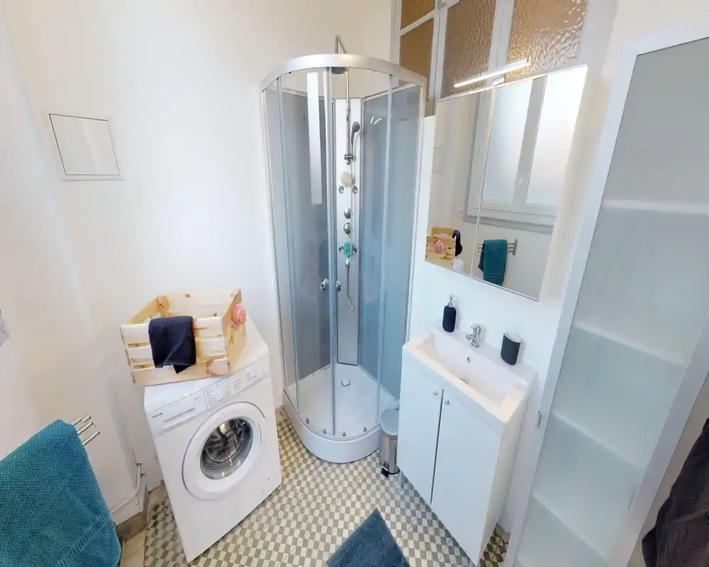 Clean, compact bathroom with a corner shower cabin, sink with mirrored cabinet, washing machine and tall storage unit; tiled patterned floor and bright natural light.