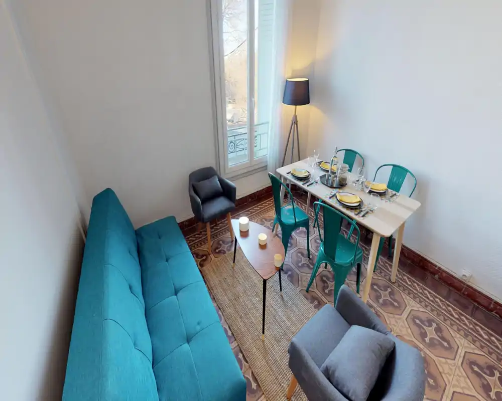 Stylish living area with a teal sofa, two armchairs, a small coffee table and a dining table set for four next to a bright window — cozy and well-styled for entertaining.