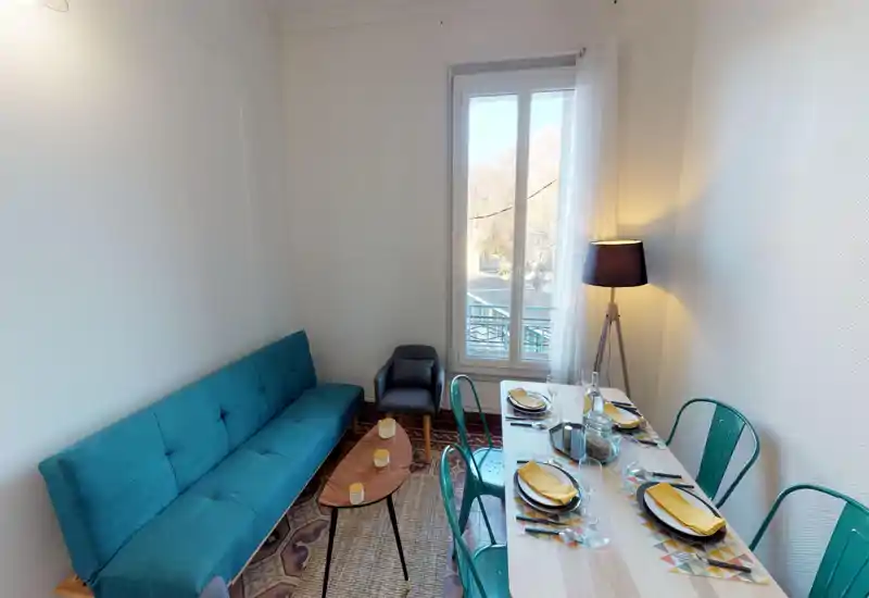 Bright, styled living area with a teal sofa, small coffee table and a dining table set for four next to a large window. The room feels cozy thanks to a floor lamp, rug and complementary accents, ideal for relaxed stays.