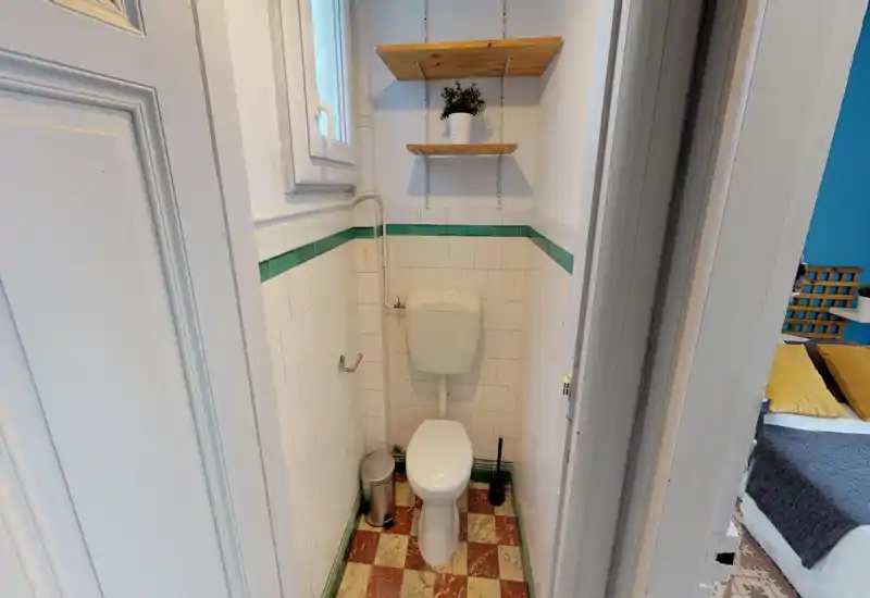 Petite salle de bain carrelée avec WC standard, réservoir mural, petite fenêtre au-dessus et étagères en bois avec une plante ; compact et propre.