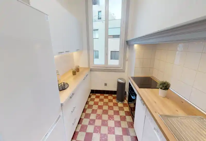 Cuisine compacte et lumineuse avec plans de travail en bois, meubles blancs, plaque de cuisson électrique et évier. Le sol à damier et la fenêtre apportent du caractère et de la lumière naturelle.