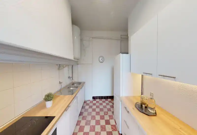 Cuisine étroite et lumineuse avec placards blancs, plans de travail en bois et sol en damier. Évier, plaque à induction et grand réfrigérateur offrent un espace de cuisson propre et fonctionnel.