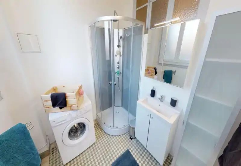 Salle de bain compacte et propre avec cabine de douche d'angle, lavabo avec armoire miroir, machine à laver et meuble de rangement haut ; sol carrelé et lumière naturelle lumineuse.