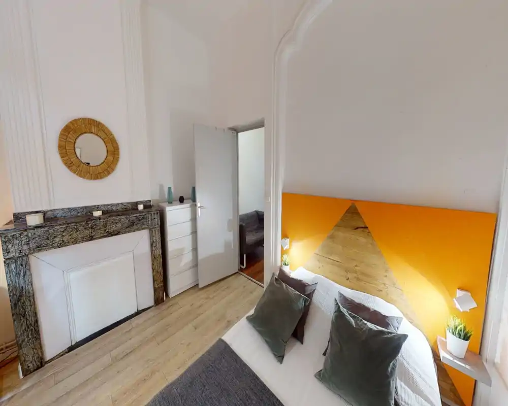 Chambre lumineuse et stylée avec une tête de lit triangulaire en bois sur un mur orange, textiles douillets et plantes de chevet. On voit un lit double confortable, une cheminée en marbre et une commode — idéal pour une location en ville.