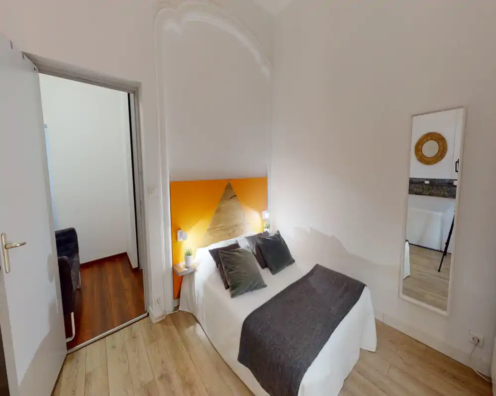 Chambre confortable et bien mise en scène avec un lit double, une tête de lit orange et bois, lampes de chevet et coussins décoratifs. Sol soigné et miroir en pied complètent cet espace lumineux et accueillant — idéal pour une annonce locative.