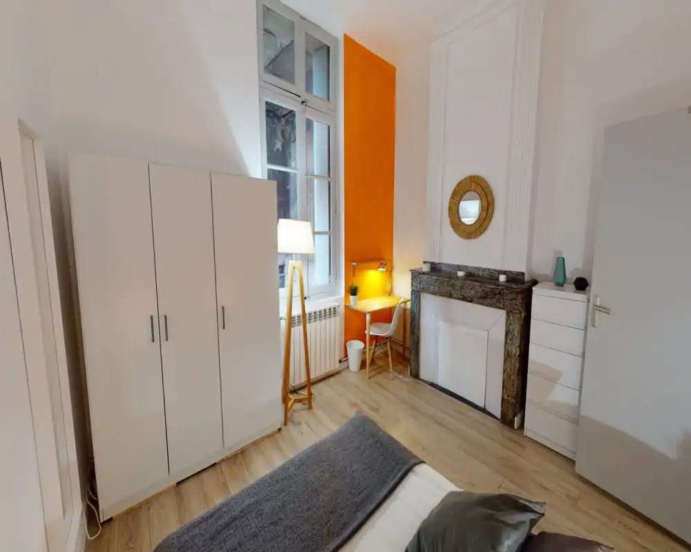 Chambre lumineuse et élégante avec lit double visible au premier plan, sol en bois, armoire blanche, petit bureau près de la fenêtre et accents décoratifs chaleureux.