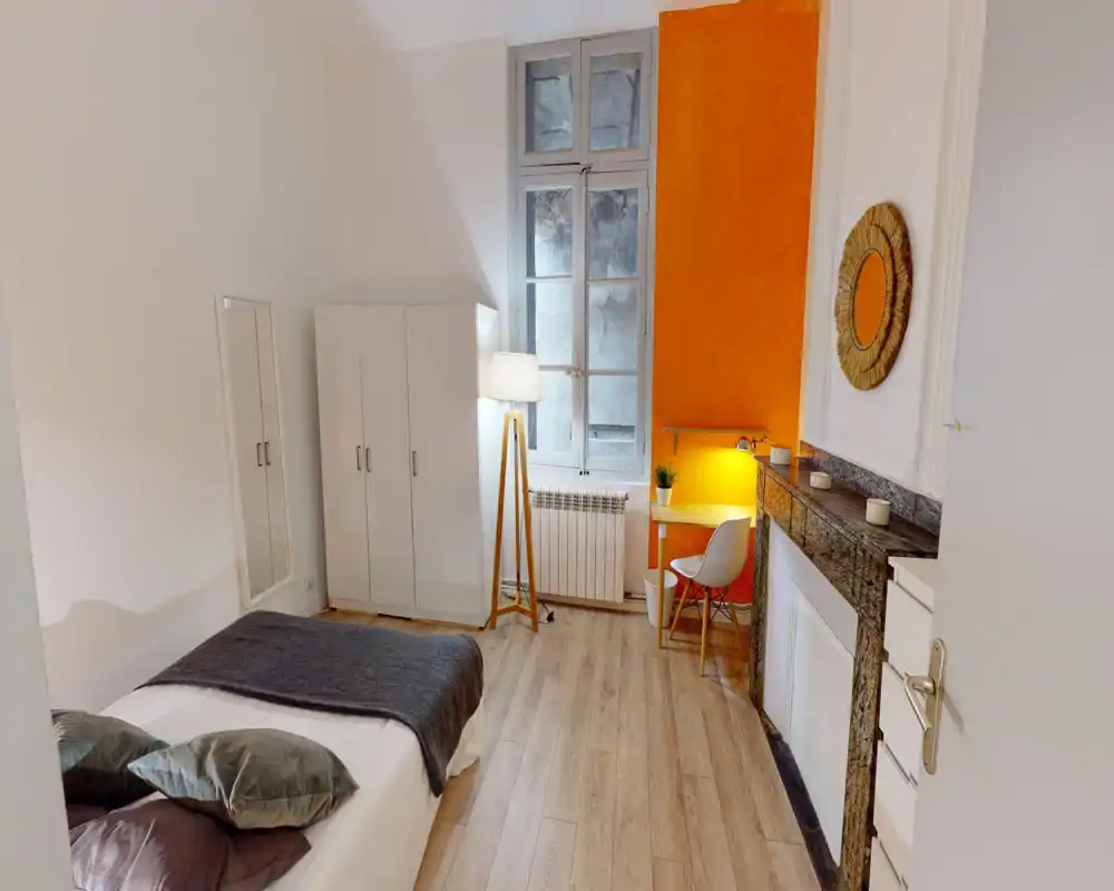 Chambre lumineuse et aménagée avec un lit double au premier plan, sol en bois, armoire et un petit bureau près de la fenêtre, mur d'accent orange et lampe chaude.
