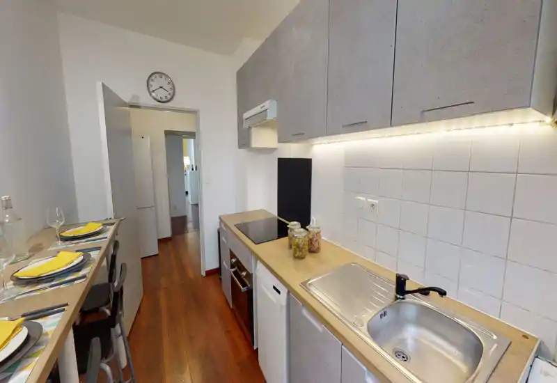 Cuisine compacte et moderne avec plan de travail en bois, évier et plaque intégrés, meubles hauts et bar petit-déjeuner dressé pour deux — lumineuse, fonctionnelle et bien mise en scène pour location.