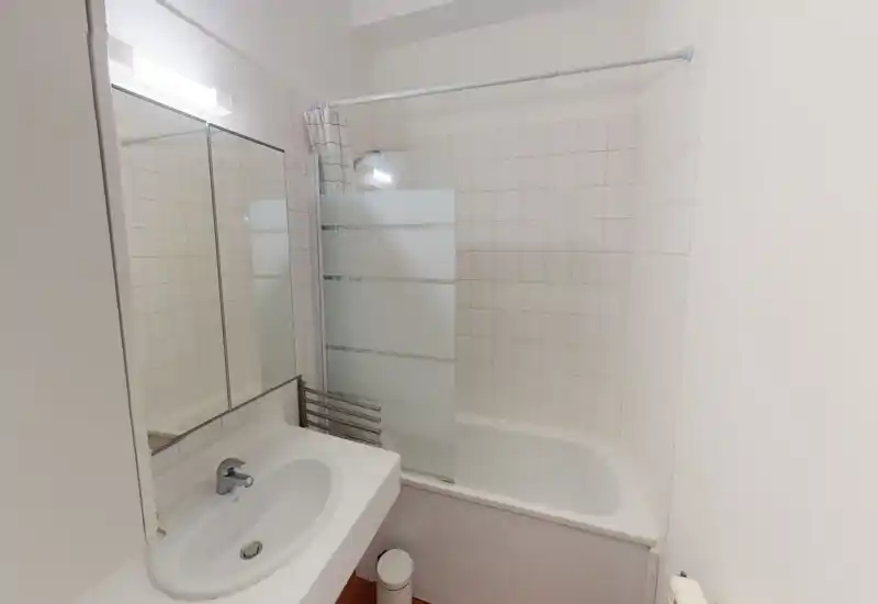 Salle de bain compacte et lumineuse avec baignoire et paroi de douche en verre dépoli, murs carrelés blancs, lavabo avec armoire miroir et robinet moderne.